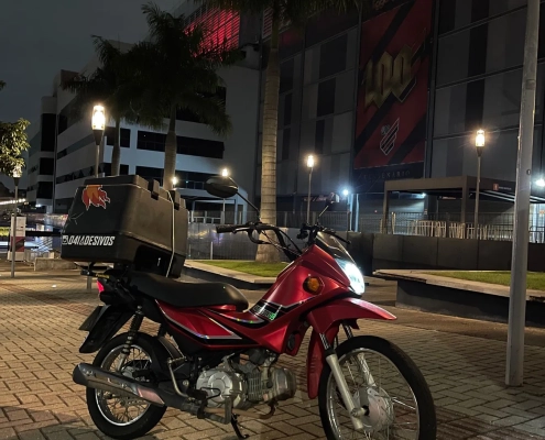 Honda Pop vermelha com baú adesivado em frente à Arena da Baixada