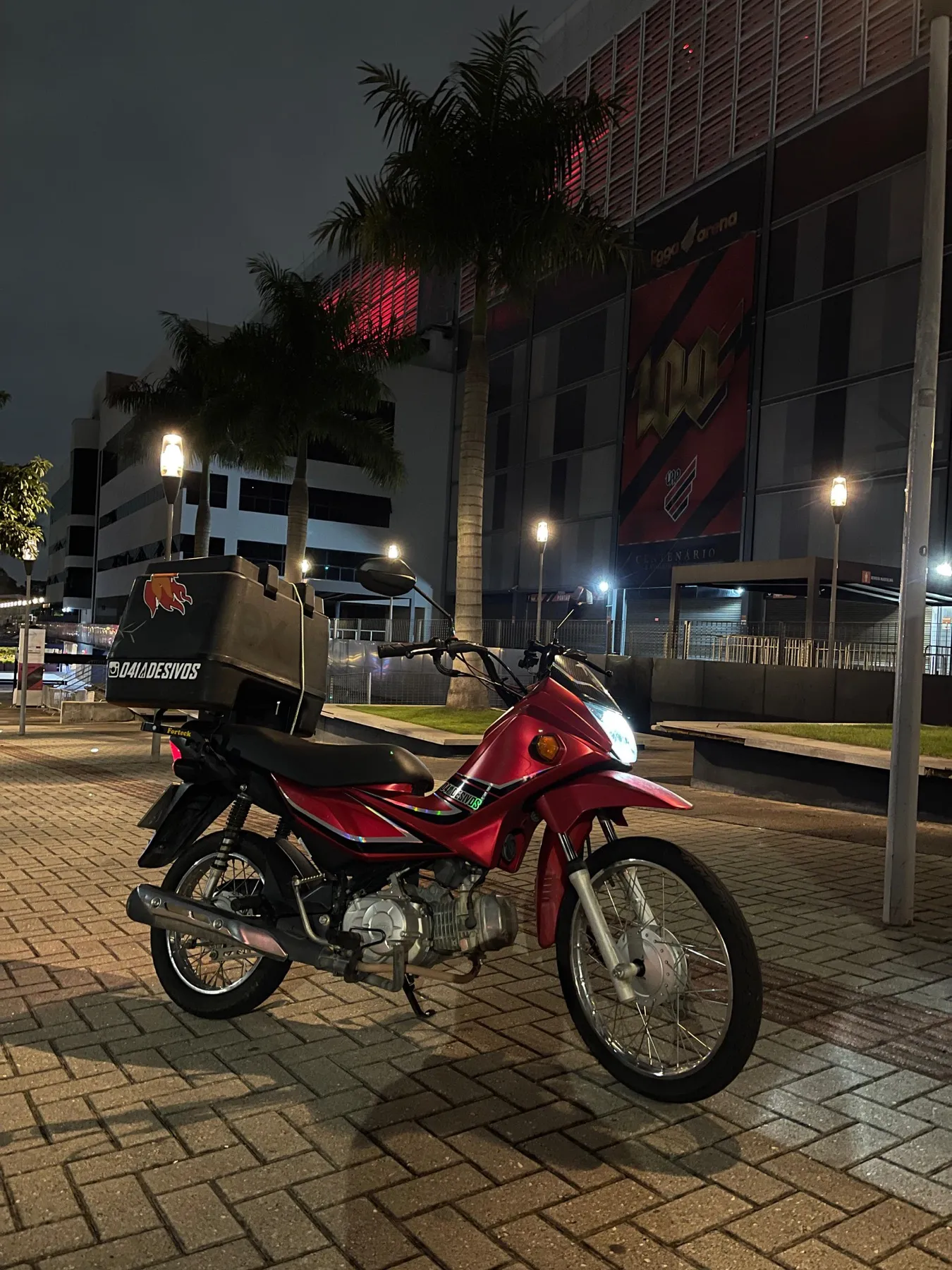 Honda Pop vermelha com baú adesivado em frente à Arena da Baixada